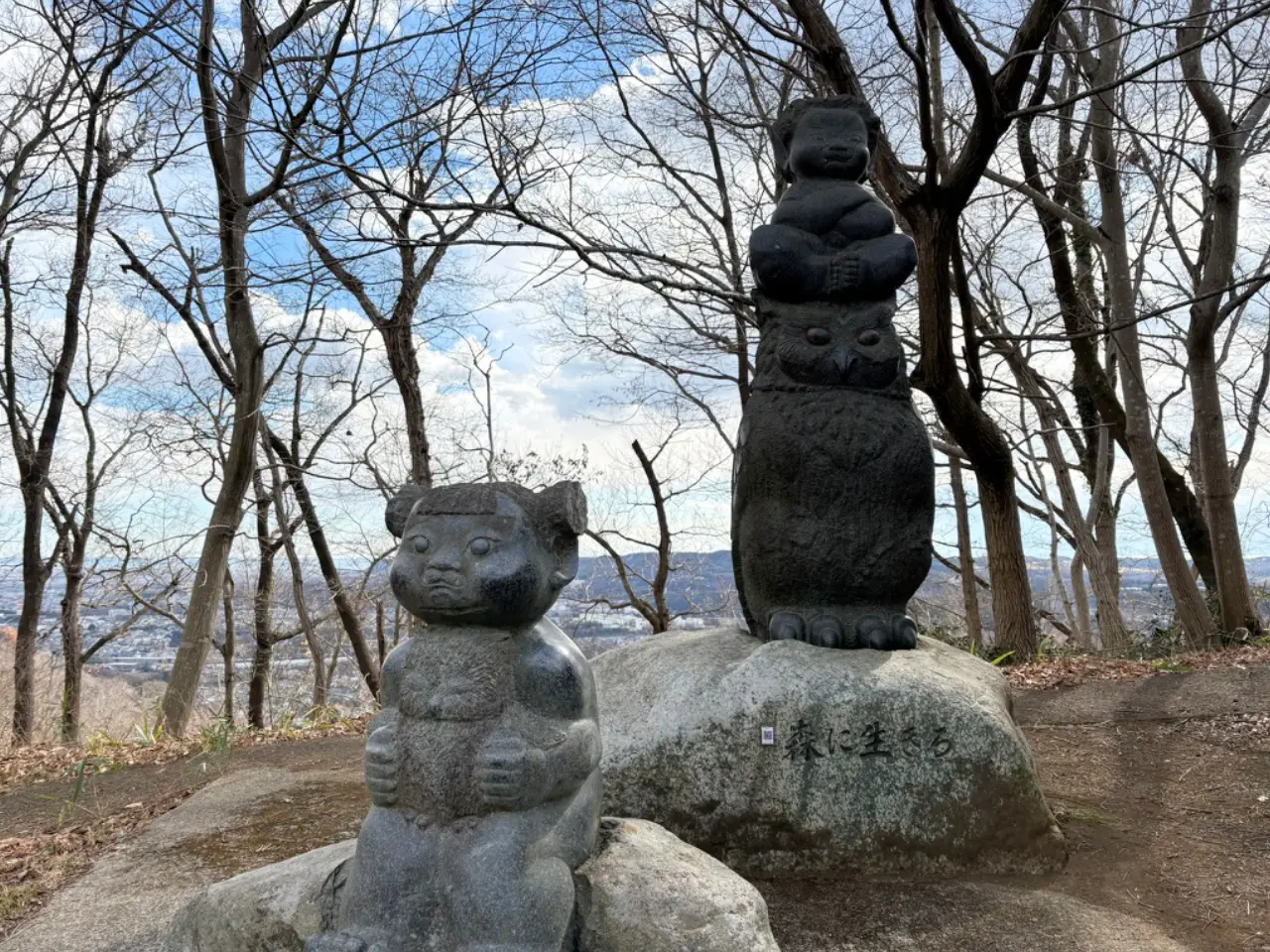 女の子がフクロウを膝にかけ、男の子が他のフクロウの頭に載っている石の彫刻。作品名は「森に生きる」